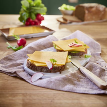 Zwei Brotscheiben mit Käse, Salat und Radieschen liegen auf einem Teller auf einem Holztisch. In einer sonnendurchfluteten Küche sind außerdem ein Messer, ein Bund Radieschen und ein Laib Brot zu sehen.