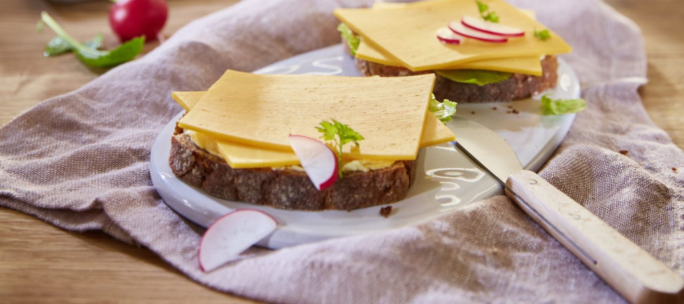 Zwei Brotscheiben mit Käse, Salat und Radieschen liegen auf einem Teller auf einem Holztisch. In einer sonnendurchfluteten Küche sind außerdem ein Messer, ein Bund Radieschen und ein Laib Brot zu sehen.