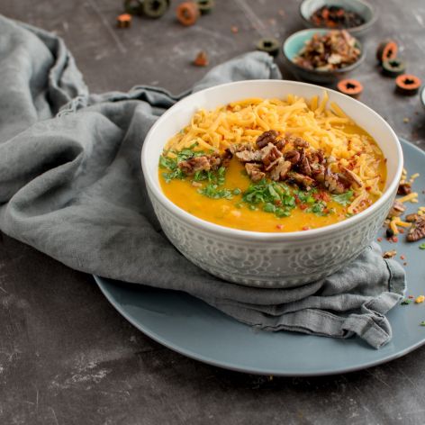 Schale mit cremiger Kürbissuppe, garniert mit geriebenem Käse, gehackten Nüssen und frischen Kräutern. Die Schale steht auf einem blauen Teller und einer grauen Stoffserviette. Rund um den Teller sind weitere Kräuter verteilt, rechts daneben liegt ein Löffel.