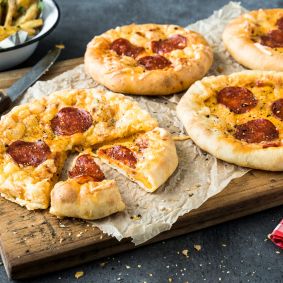 Vier Mini-American-Pizzen mit Peperoni-Belag liegen auf einem Holzbrett, eine davon angeschnitten. Eine Schüssel Pasta, ein Messer, eine Pfeffermühle und eine rotkarierte Serviette runden das Bild auf der dunklen Tischplatte ab. Perfekt für alle, die Lust auf mittelgroße Pizza haben.