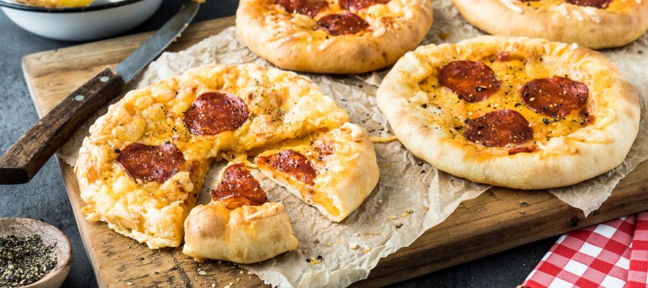 Vier Mini-American-Pizzen mit Peperoni-Belag liegen auf einem Holzbrett, eine davon angeschnitten. Eine Schüssel Pasta, ein Messer, eine Pfeffermühle und eine rotkarierte Serviette runden das Bild auf der dunklen Tischplatte ab. Perfekt für alle, die Lust auf mittelgroße Pizza haben.