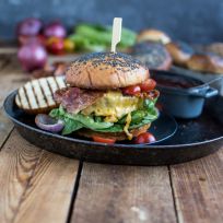 Ein Gourmet-Bacon-Burger mit Cheddar, Salat und Tomaten auf einem Mohnbrötchen liegt auf einem schwarzen Teller mit gegrilltem Gemüse und einer kleinen Schüssel Soße, alles auf einem rustikalen Holztisch.