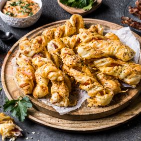 Ein hölzerner Teller mit goldbraunen, gedrehten Blätterteigstangen mit Bacon und Cheddar. Rund um den Teller liegen Zutaten wie Knoblauchzehen, Petersilienblätter, eine kleine Schale mit rotem Gewürzpulver, eine Schale mit schwarzem Pfeffer, ein Löffel sowie knuspriger Bacon im Hintergrund.