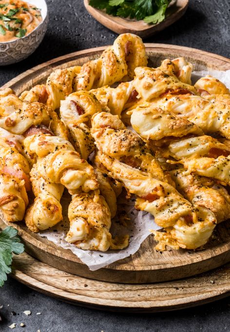 Ein hölzerner Teller mit goldbraunen, gedrehten Blätterteigstangen mit Bacon und Cheddar. Rund um den Teller liegen Zutaten wie Knoblauchzehen, Petersilienblätter, eine kleine Schale mit rotem Gewürzpulver, eine Schale mit schwarzem Pfeffer, ein Löffel sowie knuspriger Bacon im Hintergrund.