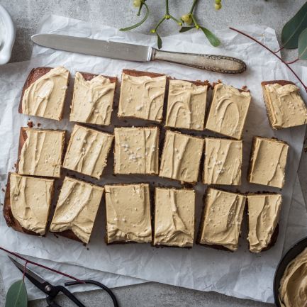 Ein rechteckiges Blech Spekulatius-Blondies mit hellbraunem Zuckerguss und weißer Schokolade wird auf Backpapier in Quadrate geschnitten. Ein Messer und eine kleine Schüssel mit Zuckerguss stehen daneben, Eukalyptusblätter liegen verstreut auf einer hellen Unterlage.