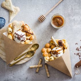 Zwei Papiertüten gefüllt mit selbstgemachten Bubble Waffles, Joghurteis, Herzkeksen, Schokoraspeln und Waffelröllchen liegen auf einem Holzbrett. Daneben liegen Karamellsauce, ein Honiglöffel, Löffel und eine blaue Serviette.