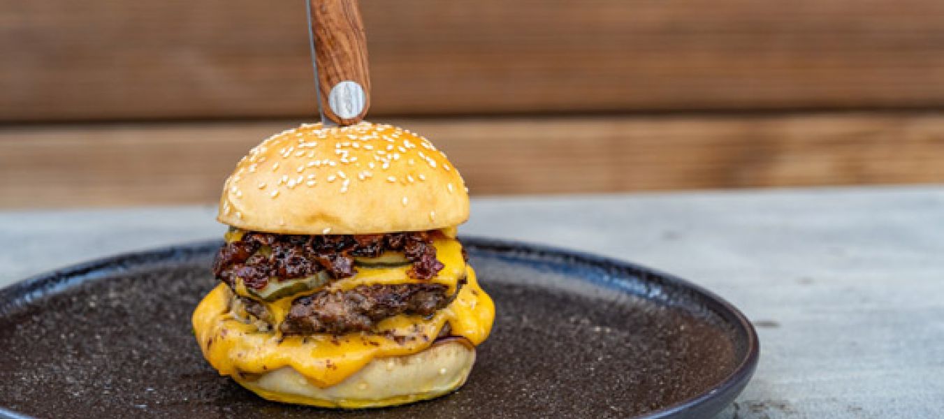 Ein Doppel-Cheeseburger mit zwei Rindfleisch-Patties, geschmolzenem Cheddar und knusprigem Speck liegt auf einem schwarzen Teller. Ein Messer steckt senkrecht in der oberen Sesambrötchenhälfte. Im Hintergrund sind Holzvertäfelungen und ein rauchiges Tomaten-Chutney zu sehen.