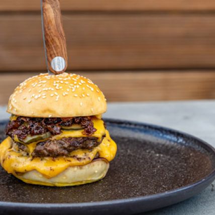 Ein Doppel-Cheeseburger mit zwei Rindfleisch-Patties, geschmolzenem Cheddar und knusprigem Speck liegt auf einem schwarzen Teller. Ein Messer steckt senkrecht in der oberen Sesambrötchenhälfte. Im Hintergrund sind Holzvertäfelungen und ein rauchiges Tomaten-Chutney zu sehen.