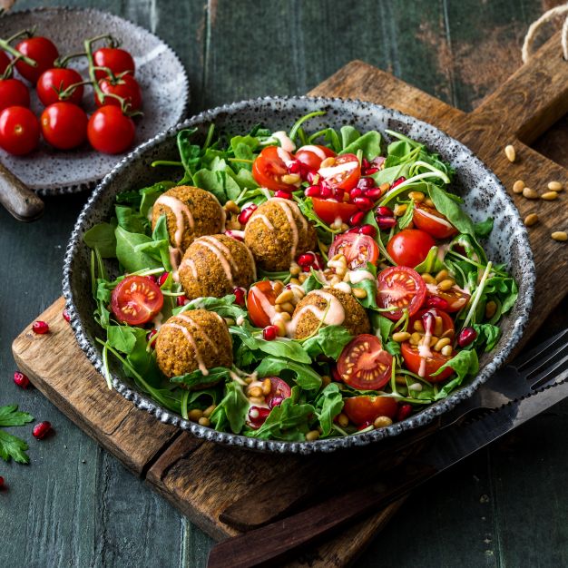 Eine Schüssel frischer Salat mit knusprigen Falafelbällchen, Kirschtomaten, Rucola, Granatapfelkernen und Pinienkernen, beträufelt mit Sesamdressing, auf einem rustikalen Holztisch mit einem Beilagenteller mit Kirschtomaten und verstreuten Zutaten.