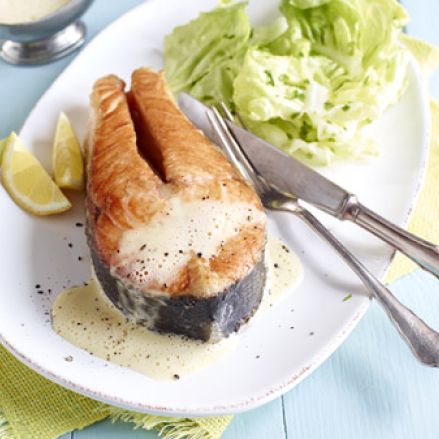 Gegrillte Lachssteaks mit cremiger Buttersauce, serviert mit Zitronenspalten und grünem Salat auf einem weißen Teller, dazu Gabel und Messer auf einem hellblauen Holztisch.