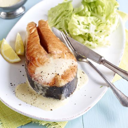 Gegrillte Lachssteaks mit cremiger Buttersauce, serviert mit Zitronenspalten und grünem Salat auf einem weißen Teller, dazu Gabel und Messer auf einem hellblauen Holztisch.