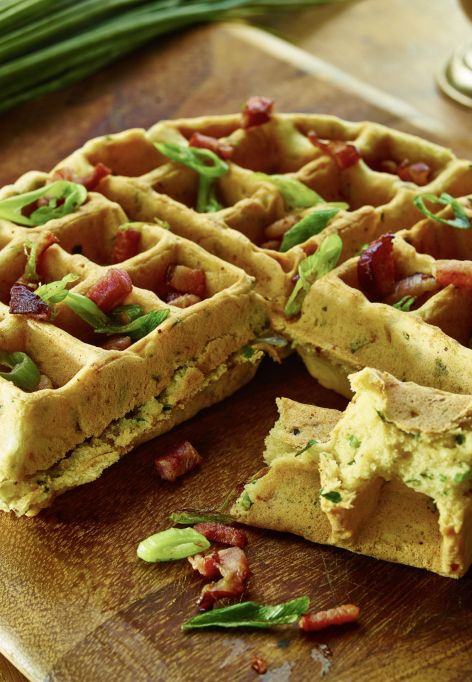 Eine Herzhafte Waffel mit gehackten Frühlingszwiebeln und Speckstücken liegt auf einem Holzschneidebrett. Daneben liegen ein Bündel frischer Schnittlauch und ein Messingbecher. Ein Stück davon wurde abgeschnitten und davor platziert.