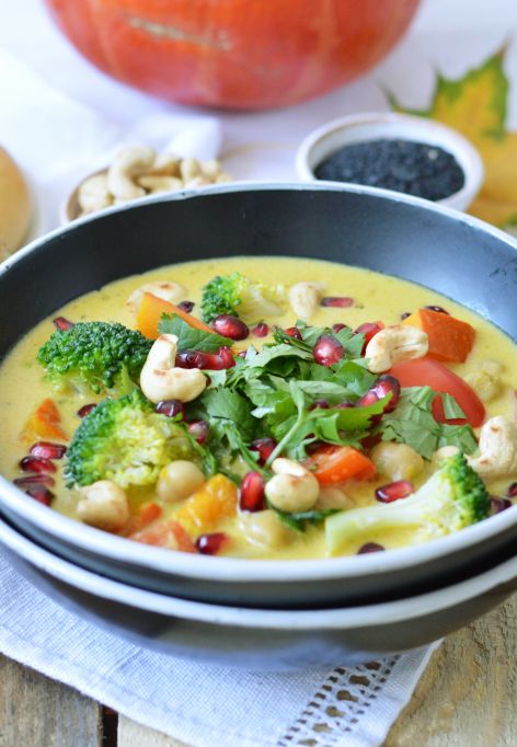 Eine Schale Kürbiscurry-Suppe, garniert mit Brokkoli, Cashewkernen, Granatapfelkernen und frischem Koriander. Die Schale steht auf einem weißen Tuch, daneben liegt ein goldener Löffel. Im Hintergrund ein Teller mit zwei Brötchen, bestreut mit schwarzem Sesam, außerdem einige Champignons, kleine Schälchen mit schwarzem Sesam und gelbem Currypulver, eine rote Chilischote und grüne Blätter.