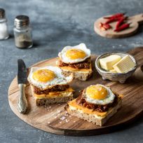 Drei belegte Brötchen auf No-Knead-Bread mit Käse, karamellisierten Zwiebeln und Spiegeleiern, serviert auf einem Holzbrett mit Speckmarmelade, einer Schüssel Butter, einem Messer, Salz- und Pfefferstreuern und roten Chilischoten im Hintergrund.