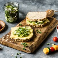 Ein rustikales Holzbrett mit einem offenen Sandwich aus Vollkornbrot, Käsescheiben, Gurkenscheiben und frischen Kräutern. Daneben steht ein Glas mit eingelegten Gurken. Links im Bild liegt ein Holzbrett mit Spießen, die mit Kirschtomaten, Gurkenscheiben und Käsewürfeln bestückt sind. Rechts steht eine weiße Schale mit Kirschtomaten, einige Tomaten und Kräuter sind locker auf dem Tisch verteilt.