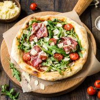 Eine frisch gebackene Pizza Rucola mit Kirschtomaten, Schinken und gehobeltem Parmesan liegt auf einem runden Holzbrett, umgeben von einem Pizzaschneider, geriebenem Käse, Salz, Pfeffer und frischem Rucola auf einem rustikalen Tisch.