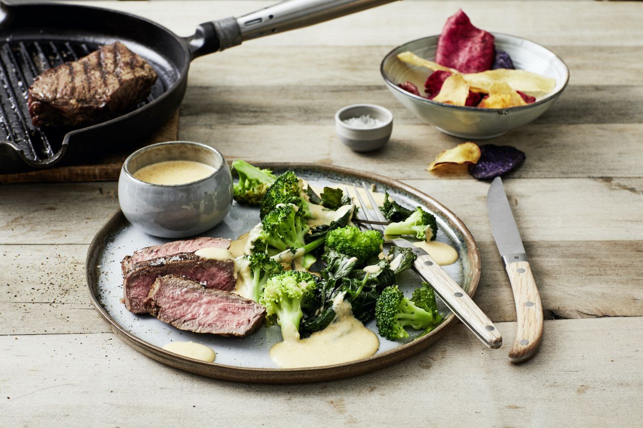 Ein Teller mit gegrilltem Rumpsteak, Brokkoli, Blattgemüse und cremiger Cheddarsauce steht neben Messer und Gabel. Im Hintergrund stehen eine kleine Schüssel mit Sauce, bunte Gemüsechips und eine Grillpfanne mit Steak.