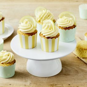 Sechs Zitronen-Cupcakes mit gewirbeltem Zuckerguss und Zitronenscheiben werden auf einem Tortenständer und einem Holztisch präsentiert, mit leeren pastellfarbenen Cupcake-Förmchen und einem angeschnittenen Cupcake auf einem Holzbrett in der Nähe.