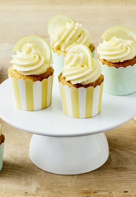 Sechs Zitronen-Cupcakes mit gewirbeltem Zuckerguss und Zitronenscheiben werden auf einem Tortenständer und einem Holztisch präsentiert, mit leeren pastellfarbenen Cupcake-Förmchen und einem angeschnittenen Cupcake auf einem Holzbrett in der Nähe.