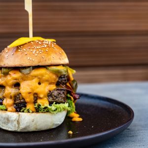 Nahaufnahme eines Cheeseburgers auf einem schwarzen Teller. Der Burger besteht aus einem Sesambrötchen, zwei Rindfleisch-Patties, geschmolzenem Käse, Gewürzgurken, Salat und einer Sauce, die an den Seiten herunterläuft. Ein Holzspieß hält den Burger zusammen.