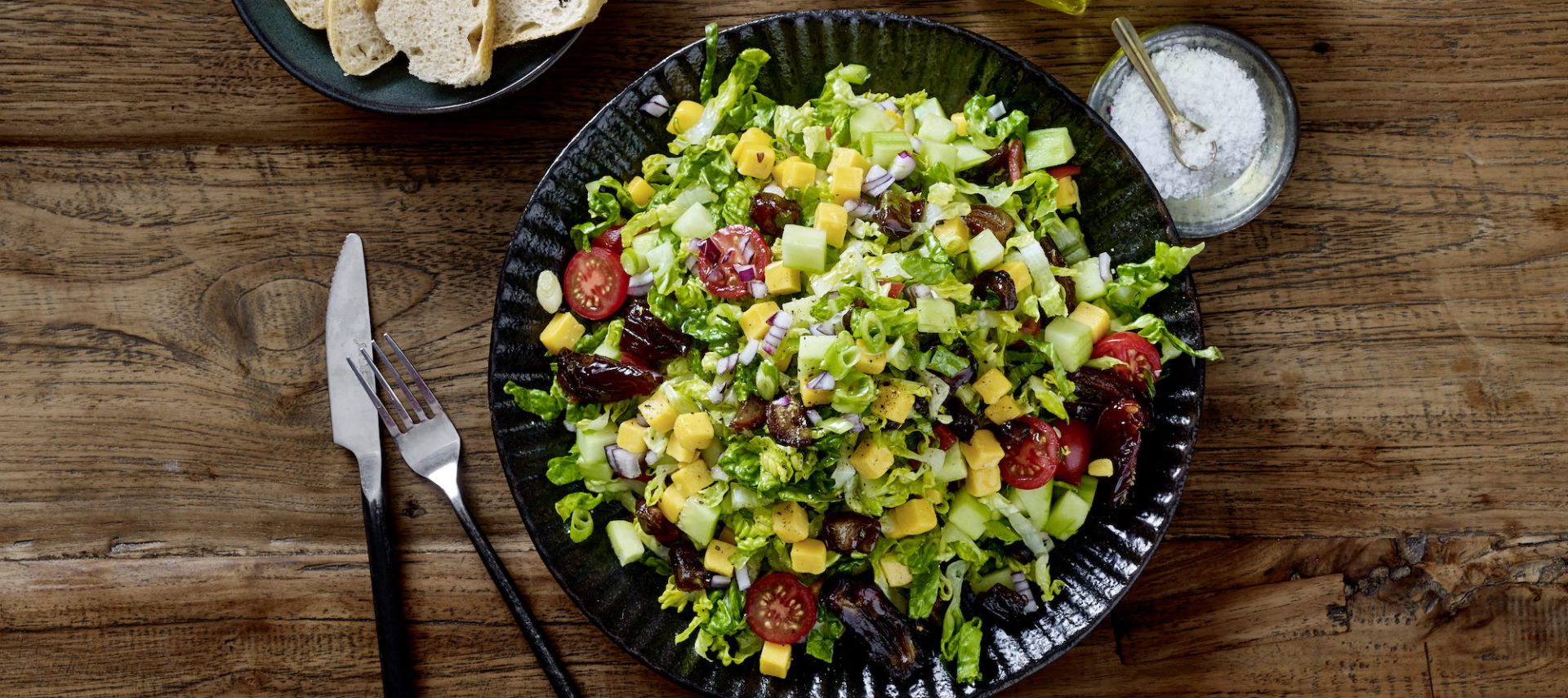 Auf einem schwarzen Teller liegt ein bunter Sommersalat mit Blattgemüse, Kirschtomaten, Gurken, Mango, Nüssen und gebratenen Datteln. Brotscheiben liegen auf einem Beilagenteller neben Olivenöl, Salz, Pfeffer und weiteren Datteln auf einem rustikalen Holztisch.