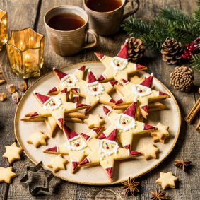Ein Teller mit sternförmigen Nikolauskeksen, die als Weihnachtsmanngesichter verziert sind, steht auf einem rustikalen Holztisch, umgeben von zwei Tassen Tee, Kerzen, Tannenzapfen, Zimtstangen und festlichem Grün.