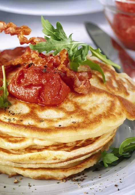 Ein Stapel herzhafter Pancakes, belegt mit knusprigem Bacon, einem Klecks Tomatensauce und Rucolablättern. Auf dem Teller liegen ein Messer und eine Gabel mit blauen Griffen. Im unscharfen Hintergrund ist eine Schale mit weiterer Tomatensauce zu erkennen.