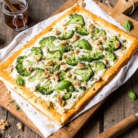 Eine rechteckige Blätterteig-Tarte, belegt mit Brokkoli, Blumenkohl, Walnüssen und frischem Basilikum, liegt auf Backpapier auf einem Holzbrett – umgeben von Kräutern, Nüssen, Pfeffer und einem kleinen Glas Honig.