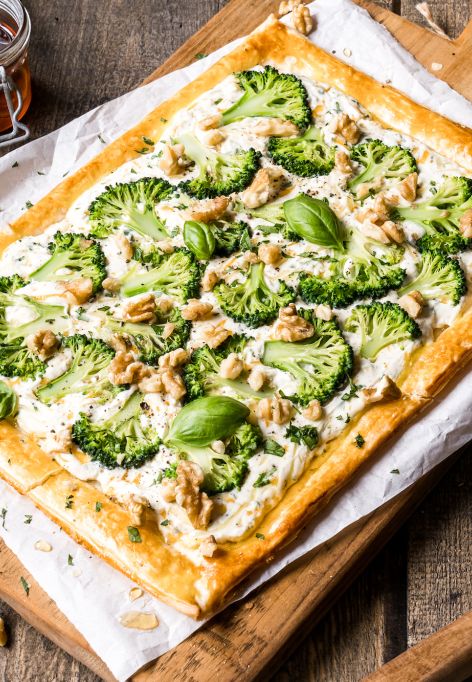 Eine rechteckige Blätterteig-Tarte, belegt mit Brokkoli, Blumenkohl, Walnüssen und frischem Basilikum, liegt auf Backpapier auf einem Holzbrett – umgeben von Kräutern, Nüssen, Pfeffer und einem kleinen Glas Honig.