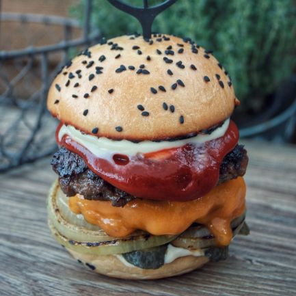 Ein von der deutschen Küche inspirierter Gourmet-Cheeseburger mit Salat, gegrillten Zwiebeln, Cheddar-Käse, Rindfleisch-Patty, Tomaten und Soßen auf einem Sesambrötchen mit einem dekorativen Zahnstocher steht auf einem rustikalen Holztisch mit Kräutern im Hintergrund.