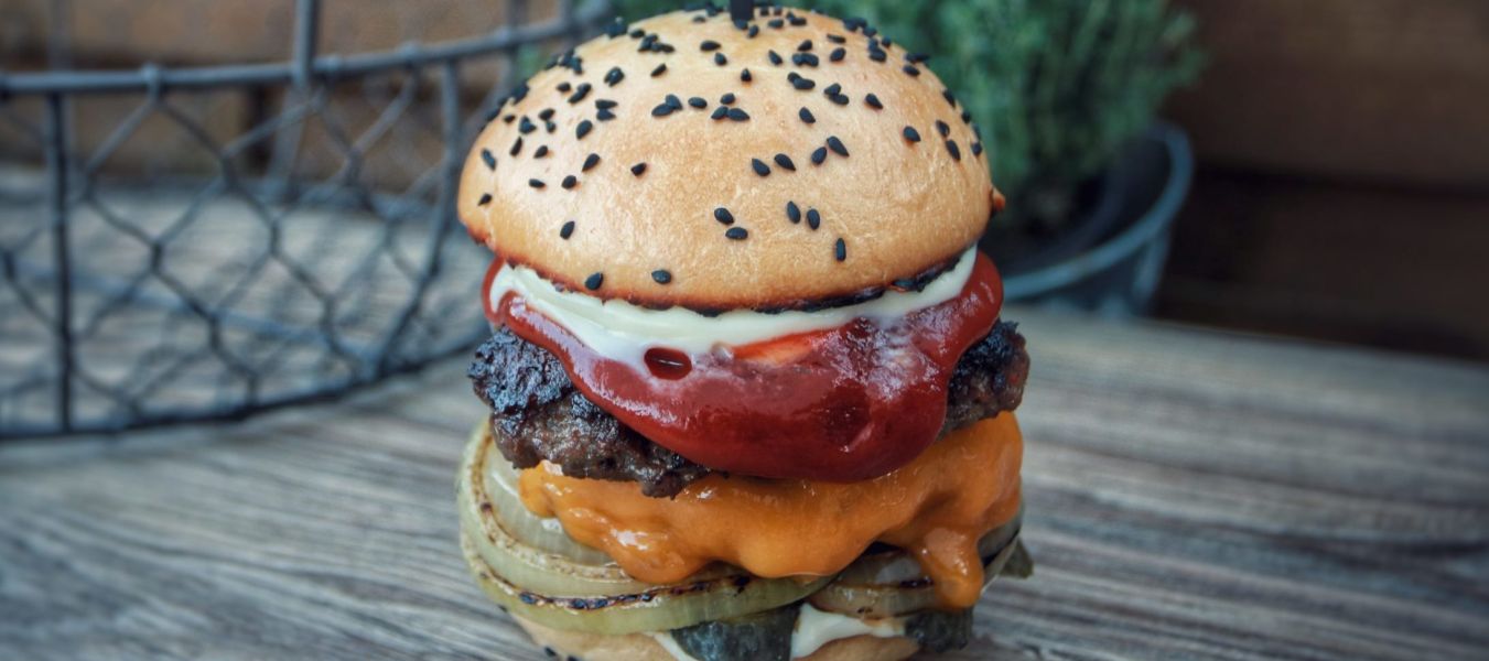 Ein von der deutschen Küche inspirierter Gourmet-Cheeseburger mit Salat, gegrillten Zwiebeln, Cheddar-Käse, Rindfleisch-Patty, Tomaten und Soßen auf einem Sesambrötchen mit einem dekorativen Zahnstocher steht auf einem rustikalen Holztisch mit Kräutern im Hintergrund.