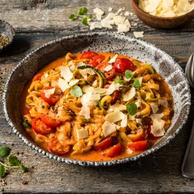 Eine Schüssel mit Zucchini-Nudeln in cremiger Tomatensauce, garniert mit Cherrytomaten, frischem Basilikum und gehobeltem Käse. Die Schüssel steht auf einem rustikalen Holztisch, daneben liegen eine Gabel und ein Löffel. Im Hintergrund sind weitere Cherrytomaten und eine kleine Schale mit Pfeffer zu sehen.