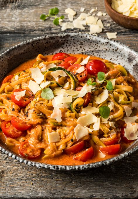 Eine Schüssel mit Zucchini-Nudeln in cremiger Tomatensauce, garniert mit Cherrytomaten, frischem Basilikum und gehobeltem Käse. Die Schüssel steht auf einem rustikalen Holztisch, daneben liegen eine Gabel und ein Löffel. Im Hintergrund sind weitere Cherrytomaten und eine kleine Schale mit Pfeffer zu sehen.