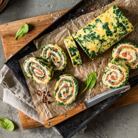 Eine Spinat-Lachs-Rolle gefüllt mit Räucherlachs und Frischkäse wird in Scheiben geschnitten und auf Pergamentpapier angerichtet, umgeben von Spinatblättern, Zitronenspalten, Chiliflocken und einem Messer auf einem rustikalen Schneidebrett.