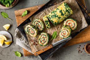 Eine Spinat-Lachs-Rolle gefüllt mit Räucherlachs und Frischkäse wird in Scheiben geschnitten und auf Pergamentpapier angerichtet, umgeben von Spinatblättern, Zitronenspalten, Chiliflocken und einem Messer auf einem rustikalen Schneidebrett.