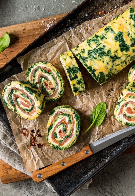 Eine Spinat-Lachs-Rolle gefüllt mit Räucherlachs und Frischkäse wird in Scheiben geschnitten und auf Pergamentpapier angerichtet, umgeben von Spinatblättern, Zitronenspalten, Chiliflocken und einem Messer auf einem rustikalen Schneidebrett.