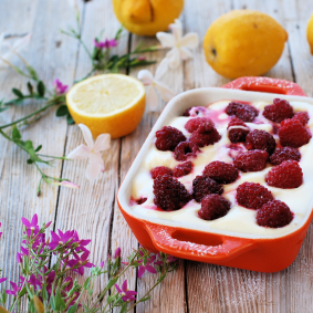 Eine rote Schale gefüllt mit cremigem Zitronen-Himbeer-Tiramisu, garniert mit Himbeeren, steht auf einem rustikalen Holztisch, umgeben von Zitronen, einer halbierten Zitrone, rosa Blumen und einem Löffel.