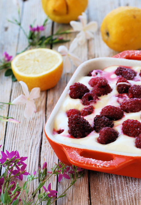 Eine rote Schale gefüllt mit cremigem Zitronen-Himbeer-Tiramisu, garniert mit Himbeeren, steht auf einem rustikalen Holztisch, umgeben von Zitronen, einer halbierten Zitrone, rosa Blumen und einem Löffel.