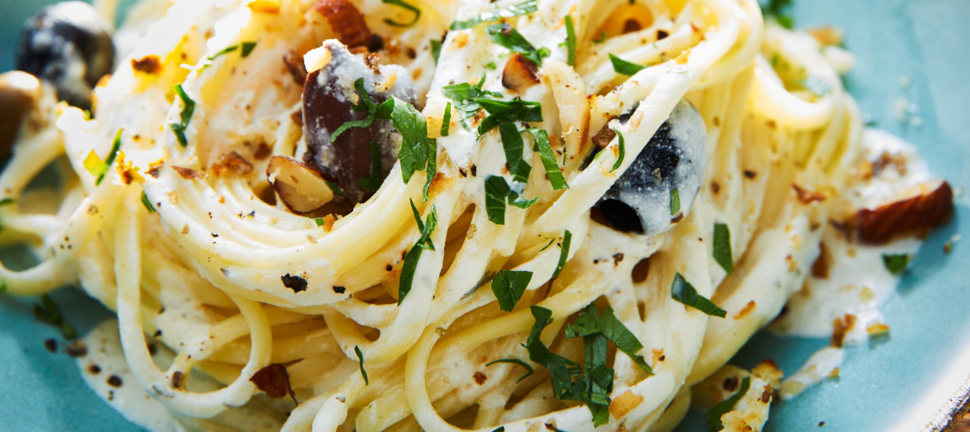 Ein Teller cremige Zitronenpasta, garniert mit frischer Petersilie, gemahlenem schwarzen Pfeffer, Oliven und gehackten Nüssen, serviert auf einer blauen Keramikschale auf einem Holztisch.