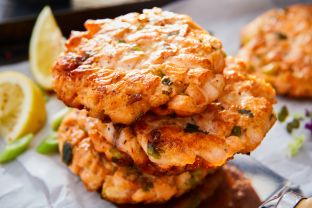 Drei goldbraune Fischfrikadellen werden auf Backpapier auf einem Backblech gestapelt, mit Kräutern garniert und mit Zitronenspalten serviert; im Hintergrund sind Salat und Teller verschwommen. Perfekt für alle, die Fischfrikadellen selbst machen möchten.