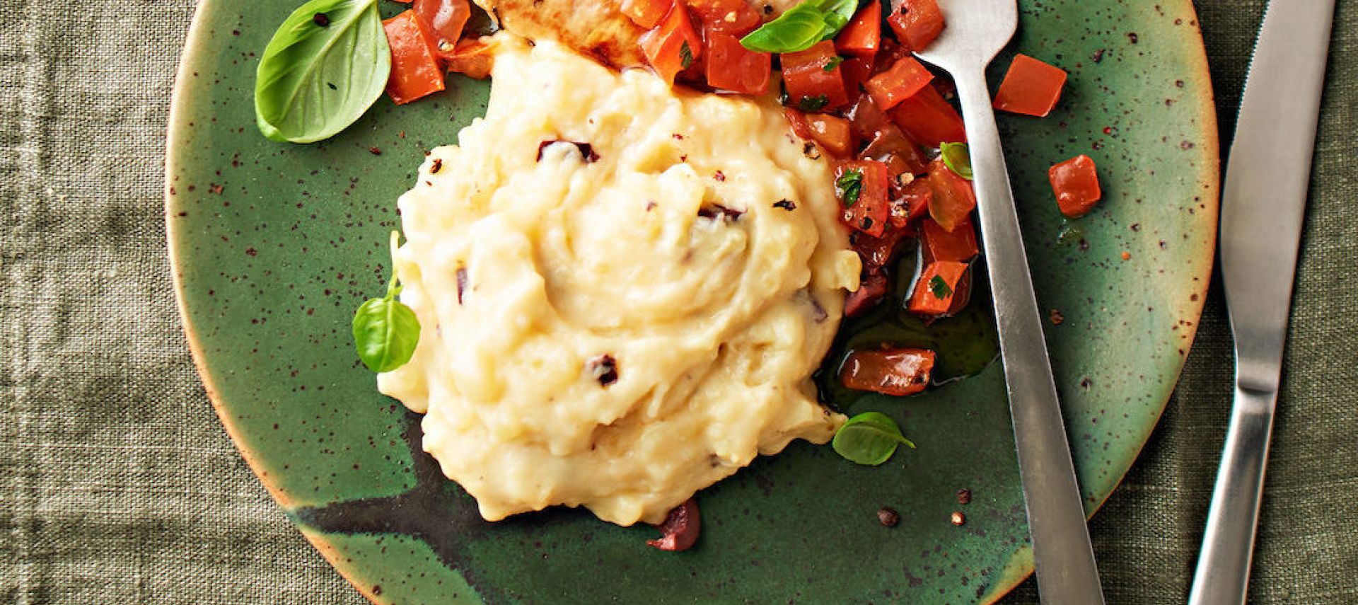 Ein grüner Teller mit würzigem Mini-Schnitzel, Kartoffel-Butter-Stampf, garniert mit gewürfelten Tomaten und frischem Basilikum. Rechts liegen Gabel und Messer, alles auf einer grünen Stoffserviette.