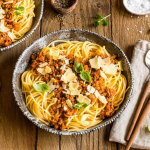 Zwei Schüsseln Spaghetti Bolognese mit Fleischsoße und gehobeltem Parmesan, garniert mit frischem Basilikum, stehen auf einem rustikalen Holztisch. Besteck, eine Serviette und kleine Schälchen mit Kräutern und Gewürzen stehen daneben.