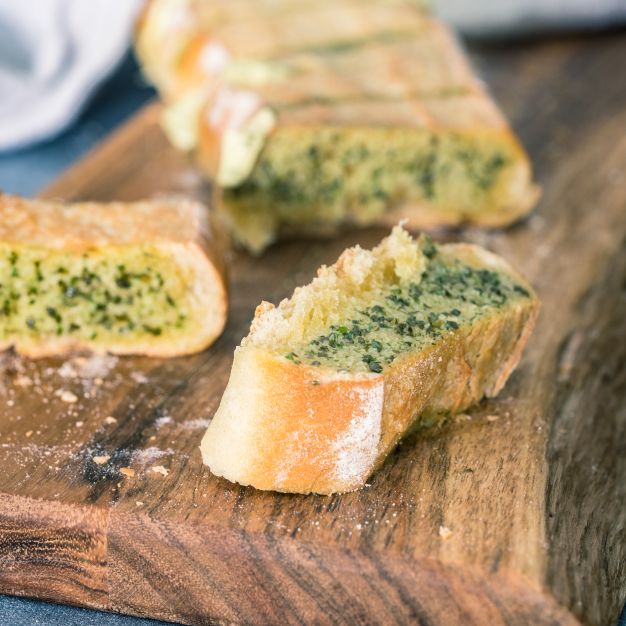 Mehrere Scheiben Kräuterbutterbaguette mit goldbrauner Kruste liegen auf einer Holzoberfläche. Die Brotscheiben sind mit einer aromatischen Kräuterbutter bestrichen.
