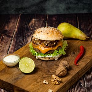 Ein Hot'n Sweet Burger mit Salat, Zwiebeln und Soße liegt auf einem Holzbrett, umgeben von einer Birnenexplosion, roten Chilis, einer halben Limette, Walnüssen, Mayonnaise und einem Keks auf einer rustikalen Holzoberfläche.