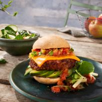 Ein Veggie-Burger mit Cheddar-Käse, Gemüse, Salsa und Tomatenmarmelade auf einem Brötchen liegt auf einem Teller neben einer Salatschüssel auf einem rustikalen Holztisch mit Äpfeln und Wassergläsern im Hintergrund.