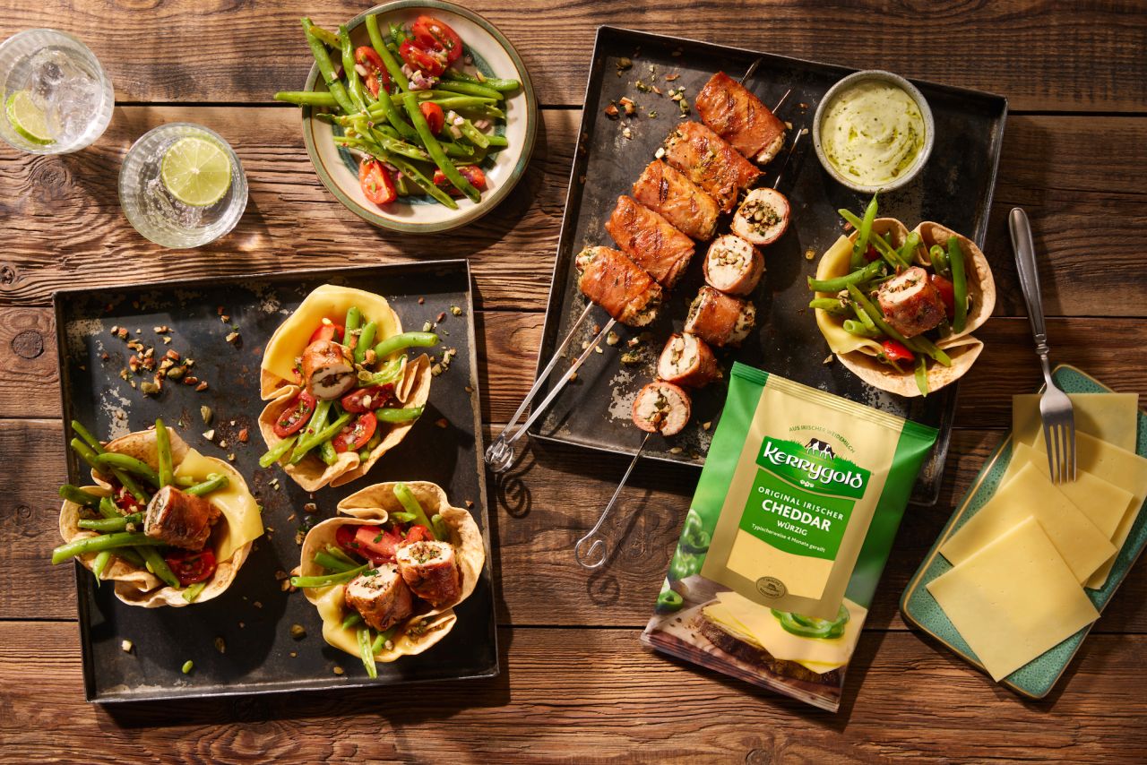 Draufsicht auf Teller mit gegrillten Hähnchenspießen, Salat und BBQ-Taco-Bechern gefüllt mit Hähnchen, Käse, grünen Bohnen und Tomaten. Eine Packung Kerrygold-Cheddar-Käsescheiben und Limettengetränke stehen ebenfalls auf dem Holztisch.