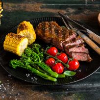 Ein schwarzer Teller mit geschnittenem Steak, gegrilltem Maiskolben, Broccolini und gerösteten Kirschtomaten. Messer und Gabel liegen auf dem Teller. Wellenförmige Pommes Frites und eine Schüssel mit grobem Salz stehen daneben auf einem rustikalen Tisch.