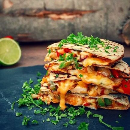 Ein Stapel käsiger Gemüse-Quesadillas mit geschmolzenem Käse und gehackten Kräutern obendrauf, serviert auf einer dunklen Schieferplatte mit frischen grünen Kräutern und einer halbierten Limette im Hintergrund – perfekt für Ihr nächstes Treffen auf bbqpit.de am 22. Juli.