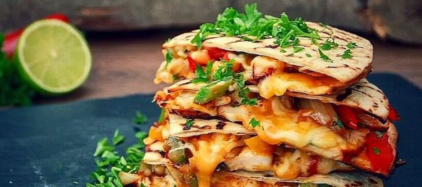 Ein Stapel käsiger Gemüse-Quesadillas mit geschmolzenem Käse und gehackten Kräutern obendrauf, serviert auf einer dunklen Schieferplatte mit frischen grünen Kräutern und einer halbierten Limette im Hintergrund – perfekt für Ihr nächstes Treffen auf bbqpit.de am 22. Juli.