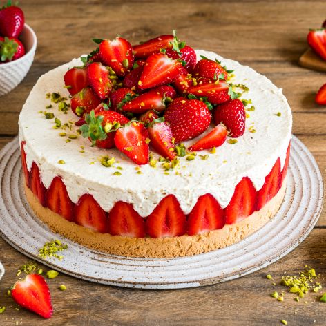 Ein Erdbeerkuchen, inspiriert von der klassischen deutschen Erdbeertorte, ist mit frischen Erdbeeren und gehackten Pistazien belegt. Geschnittene Erdbeeren säumen den Rand des Kuchens, weitere ganze Beeren und Pistazien liegen verstreut auf dem Holztisch daneben.